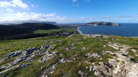 A view from the top of a large hill, the little Orme, looking out over a large crescent bay. At the other end is another large hill of the great Orme, jutting out into the ocean. The sea is a mostly calm, mid blue, The sky fades from bright to a pale blue with dotted fluffy white clouds in the distance.
