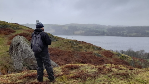 A walker standing on a hillside with rocky outcrops, grass and brash areas. They are looking out at Bala lake. The water is calm, the shore opposite is faded in a slight mist and the sky is grey.