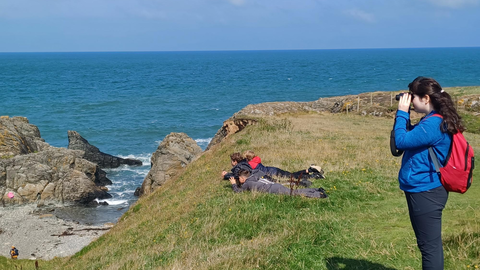 A girl stood on a grassy cliff facing left with binoculars looking down into a cove. Behind her, 3 boys are lying down near the cliff slope on their fronts to get a better view. A small cove with stony beach is visible to the left below them, and a wide blue sea behind them.