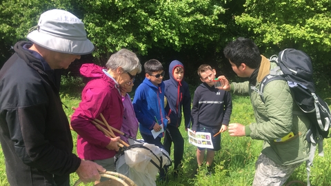 A Group with adults and young people in a field lined with trees. They are holding bug hunting nets, ID guides and bug pots. The group are gathered round looking at the insect caught in one bug pot.