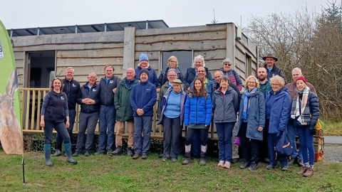 A group of 22 volunteers and staff, wearing mainly blue fleeces or waterproofs, stood on the grass outside a wooden single level building.