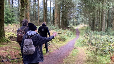 People walking down a path through the trees. The woodland ahead of them is mainly coniferous and quite dense. The person closest is pointing to the right at something of interest they have seen.