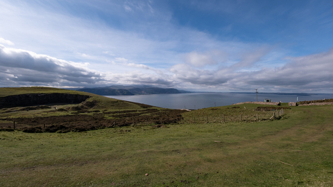 A view from the Great Orme, Grass and heathland hills drop off down to the sea. In the distance the mainland curves around enclosing the ocean like a bay. Above clouds are gathered in rows, fluffy and dark, with a deep blue sky.