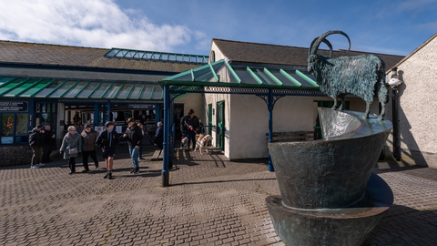 A large building with metal and glass roof verandas, brick paving outside, and a metal goat sculpture. There is a group of people and dogs leaving the building on a clear sunny day.