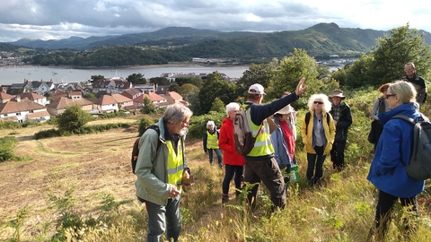A group of 10 people, some in high vis stood near the top of a sloping field. Below them is a town and sea inlet, with large hills rising on the opposite shore.