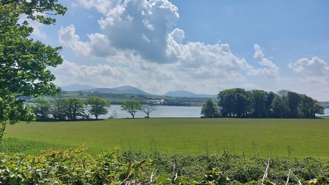 A field surrounded by hedgerows and trees. Beyond it lies the Menai Straits, and in the distance the mountains of Snowdonia and a blue sky with fluffy clouds.