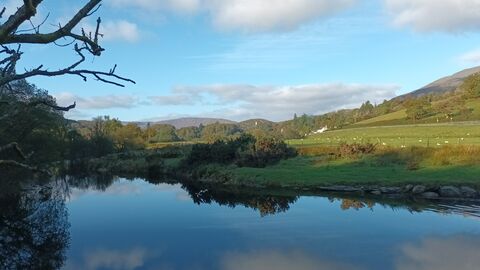 The Lledr river, a calm wide river, with trees on one bank and gently sloping farm fields leading up to a hill on the other bank. The river is dark in shadow, but with clouds and blue sky reflected in it.