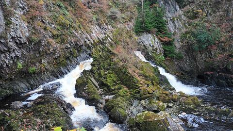 Sheer rocky river banks meet in a series of small waterfalls and rapids. The river splits in 3 directions around large rocky outcrops, with vegetation, evergreen and bare branched trees of winter clinging to the slopes.