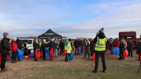 A large gathering of people in winter clothing, in a sandy dune edge car park. Most are holding litter pickers and blue or red bags, one at the front is wearing high visibility yellow and addressing the crowd through a megaphone. The day is clear and cold with a pale blue sky and light streaky clouds.