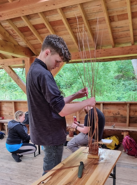 Trainee making willow bird feeder by weaving willow in an outdoor classroom