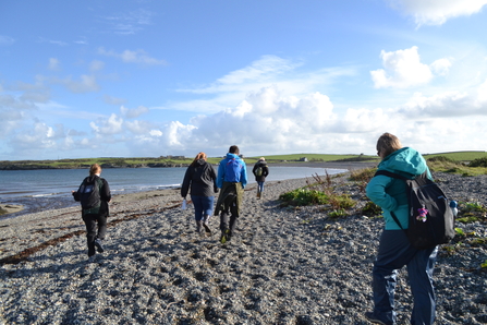 5 young people walking away from the camera, along the shingle at Cemlyn Bay dressed warmly on a sunny day