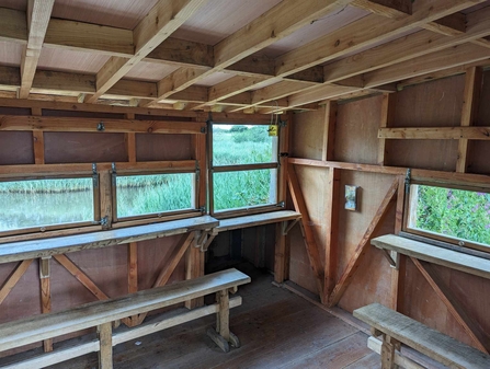 Photo of the inside of the Viley Hide at the Spinnies Aberogwen Reserve. There are two benches to sit on and several viewing windows out into the lagoon.