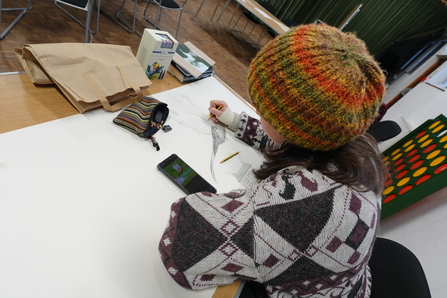 A person wearing a colourful knitted hat sketching a detailed illustration of a pheasant on a large piece of paper, using a phone image for reference, at a table in a community hall