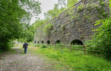 A man is walking on a wide stone path in front of a stone kiln structure, originally built in the 1850's. A bank of six archways can be seen along the front of this top draw kiln