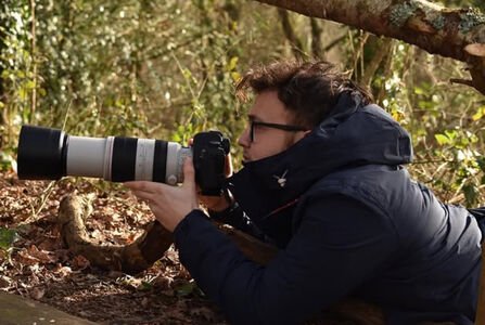 Young white man with glasses lying in a forest with a camera and a long telephoto lens