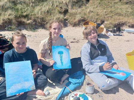 three young people holding marine themed paintings on the beach