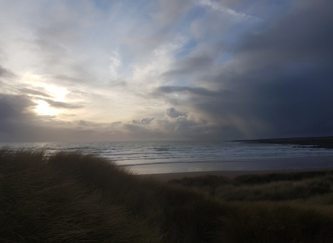 A beach with sand dunes and marram grasses at the front, in shadow. A headland curves around the right side of the beach, and there are lots of small waves coming towards shore. The sky is full of clouds, from very dark on the right, to pale and fluffy further left where the sun is breaking through with some patches of blue above them. The sun is very low in the sky and gives off a cold light that makes it obvious it is winter.