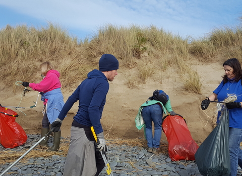 A group of 5 people in brightly coloured clothing, carrying litter pickers and bright red rubbish bags, collecting plastic and other waste from a pebbly area of a beach, in front of some sand dunes.