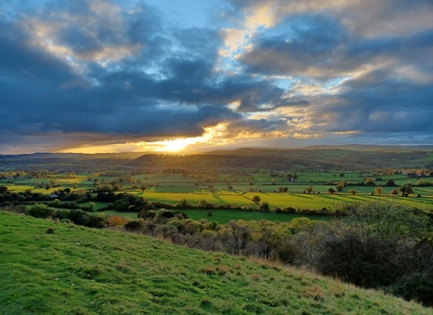 The view from Y Graig Nature Reserve across a patchwork of farm fields and distant hills. The sky is blue but clouded, with bright evening sun breaking through just above the horizon, casting long shadows and tinting the landscape with a golden glow.