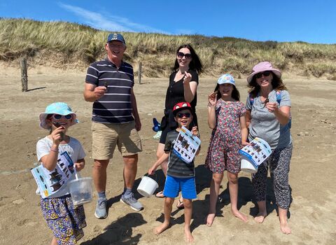 A group of 3 adults and 3 children on a sandy beach. Each is holding up an egg case or mermaids purse, The children also have buckets and ID guides. Behind them is a grassy sand dune and bright blue sky.