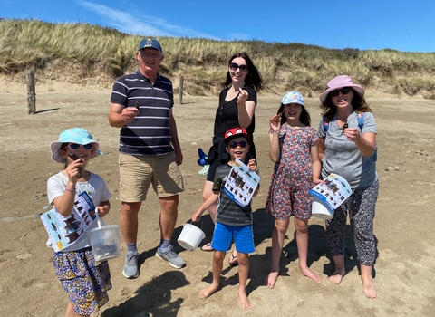 A group of 3 adults and 3 children on a sandy beach. Each is holding up an egg case or mermaids purse, The children also have buckets and ID guides. Behind them is a grassy sand dune and bright blue sky.