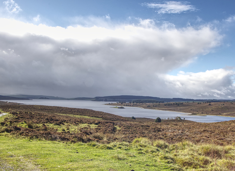 A large lake surrounded by moorland, mainly brown with dried up heather, but some green grasses in the foreground and fields to the left. A small track and fence line separates the  fields from the reserve. On the far side of the lake hills rise to enclose the area, with a shadow cast over them by immense fluffy clouds. Above them the sky is a deep blue, and the sun is illuminating the cloud edge so it glows bright white.