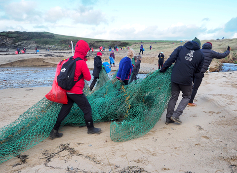 A group of 7 people hauling a washed up fishing net off a beach. In the background many more people can be seen litter picking during a very large beach clean at plastoff 2022