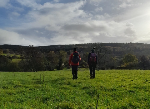 2 people in outdoor clothing, woolly hats and waterproofs, walking through an open field towards a hilly landscape with lots of tree cover.