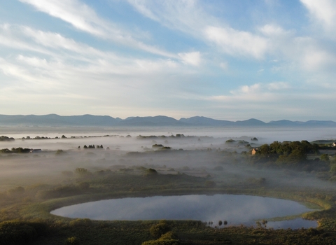 Scenic view of Cors Goch Nature Reserve on Anglesey, showcasing a serene landscape with misty wetlands, a reflective pond in the foreground, scattered trees, and distant mountains under a sky filled with soft clouds. The image captures the tranquility and natural beauty of the reserve